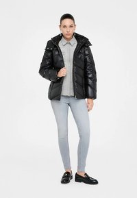 Fekete puffertöltéses dzseki magas gallérral, fényes textúrával, elülső cipzárral és patentokkal a vállakon, világosszürke ing és farmer fölött viselve.