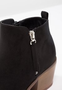 Bottine en suède noir avec fermeture éclair latérale argentée et languette de traction, dotée d'un talon en bois empilé.