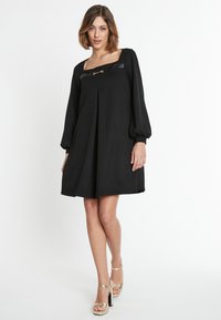 Schwarzes Kleid mit quadratischem Ausschnitt, plissiertem Rock und langen Puffärmeln. Hat ein Fake-Leder-Element und eine goldfarbene Akzentuierung.
