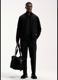 Juoda bombonešio striukė dėvima ant juodos marškinėlių, derinama su juodomis kelnėmis ir batais. Laiko juodą sportinį krepšį su užtrauktuku ir dirželiais.