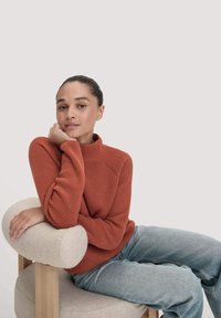Junge Frau mit zurückgebundenem Haar, die einen rostfarbenen gestrickten Pullover und hellblaue Jeans trägt, entspannt auf einem beigefarbenen gepolsterten Holzstuhl sitzt.