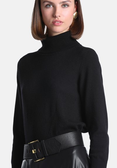 Jeune femme aux cheveux bruns courts portant des boucles d'oreilles dorées, un pull noir à col roulé et une large ceinture en cuir noire avec une boucle dorée.
