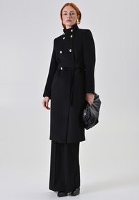 Zwarte jas met dubbele rijgouden knopen, hoge kraag, strikceintuur en slanke silhouet. Gecombineerd met zwarte wijde broek en tas.