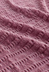 Teksturowana tkanina w kolorze mauve z wypukłymi wzorami geometrycznymi, prezentująca miękki i puszysty wygląd, złożona w taki sposób, aby ukazać głębię i detal.