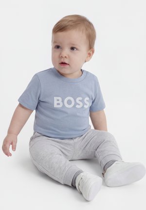 Småbarn siddende iført en lyseblå skjorte med teksten "BOSS", grå bukser og hvide sko, der kigger let til siden på en hvid baggrund.