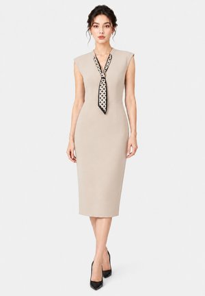 Vrouw draagt een beige mouwloze midijurk met een patroon halslint en zwarte hoge hakken, staand tegen een effen lichte achtergrond.
