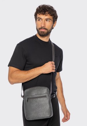 Homme barbu portant un t-shirt noir et un pantalon tenant la sangle d'un sac en cuir gris en bandoulière sur un fond uni.