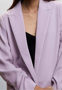 Blazer violet clair à la coupe ajustée, avec des revers crantés et des manches longues, porté sur un haut noir, présentant une texture lisse.