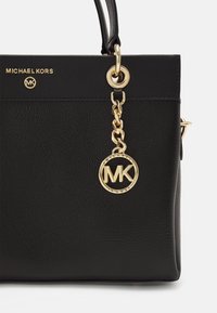 Sac en cuir noir Michael Kors avec chaîne dorée et breloque circulaire du logo MK suspendue près de l'attache de la poignée.