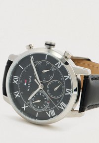 Ανδρικό ρολόι Tommy Hilfiger με μαύρη δερμάτινη λουρί, μαύρο καντράν, ασημένια θηκή και ρωμαϊκούς αριθμούς που δείχνουν ημέρα, ημερομηνία και 24-ωρη ώρα.