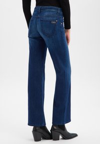 Jean en denim bleu foncé à jambes larges avec poches arrière, coutures discrètes et petit logo en accent. Associé à des bottines à talons noires.