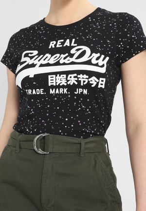 Black cotton t-shirt with star pattern, featuring a white “SuperDry” logo. Paired with olive-green pants and a silver crescent buckle.