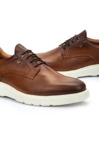 Chaussures en cuir marron avec une finition lisse, dotées de lacets noirs, d'un bout rond et d'une semelle en caoutchouc blanc contrastante. Petit logo sur le côté.