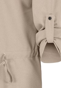 Tissu beige avec une manche retroussée fixée par une sangle boutonnée et un détail de ceinture à la taille sur un vêtement.