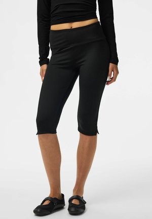Femme portant des leggings noirs courts et des chaussures style Mary Jane noires, debout devant un fond uni.