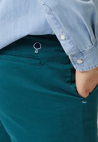 Chinos verde azulado con una textura suave, que cuentan con un bolsillo trasero y un logo de árbol bordado en azul marino. Combinados con una camisa de botones azul claro.