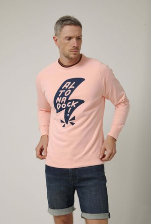Hombre con sudadera rosa con un gráfico negro de un rayo y el texto "¡ALTONA DOCK!", lleva pantalones cortos de mezclilla oscura remangados, de pie contra un fondo liso.