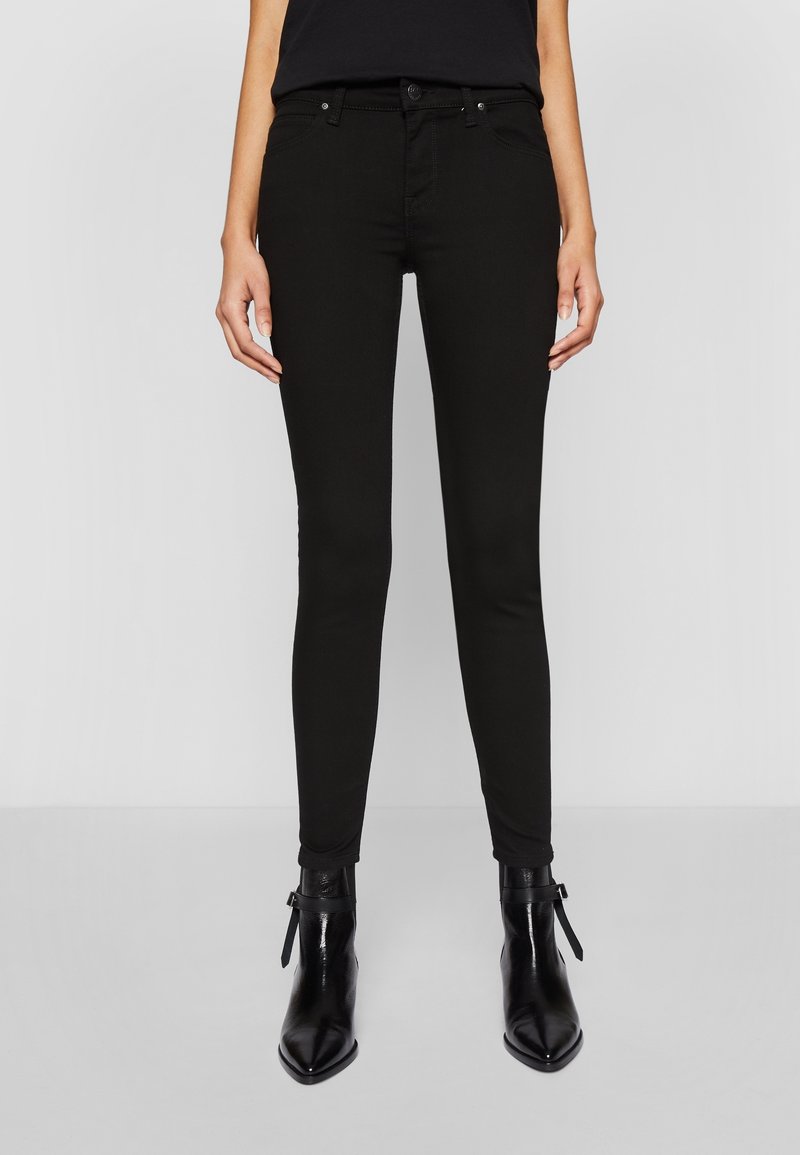 Jean skinny noir, tissu en mélange de coton, taille mi-haute, cinq poches, passants de ceinture, coupe ajustée jusqu'à la cheville, associé à des bottines noires.