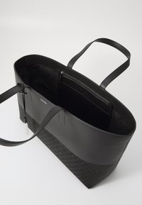 Černá taška přes rameno s hladkým koženým vrškem a texturovaným tkaninovým dnem, zdobená vzorem loga. Obsahuje dva popruhy a vnitřní kapsu na zip.