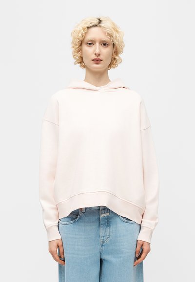 Blonde Person mit kurzen lockigen Haaren, die einen hellrosa übergroßen Hoodie und blaue Jeans trägt, steht vor einem schlichten hellen Hintergrund.