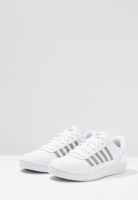Scarpe da ginnastica bianche con una tomaia in materiale sintetico liscio, caratterizzate da tre strisce grigio metallizzato, punta rotonda e suola testurizzata per una migliore aderenza.