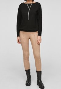 Zwarte getextureerde top met lange mouwen en een ronde hals, gecombineerd met strakke beige suèdeachtige broeken en zwarte enkellaarsjes.