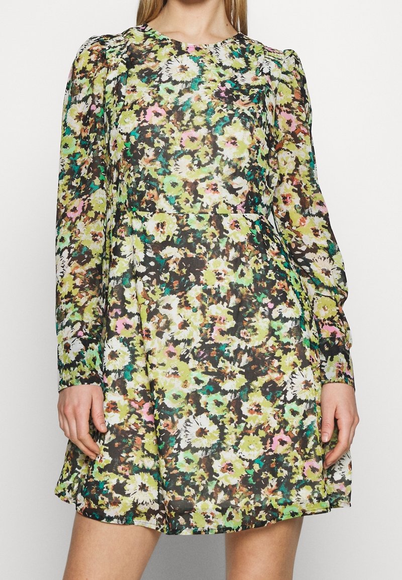 Femme portant une robe à manches longues, au-dessus du genou, avec un imprimé floral coloré vert, jaune, rose et blanc, debout sur un fond uni.