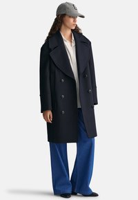 Granatowy dwurzędowy płaszcz z dużymi klapami, w połączeniu z jasną bluzką w paski i szerokimi niebieskimi dżinsami. Szary kapelusz dopełnia stylizację.