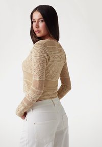 Knit beige crop top featuring intricate patterns and long sleeves. Paired with white high-waisted pants, showing a fitted silhouette.