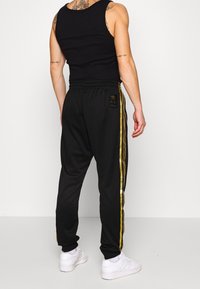 Svarta träningsbyxor med guldiga sidostråk, med elastisk midja och ribbade muddar, kombinerat med en svart linne. Vita sneakers är synliga.