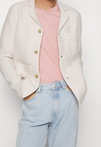 Jasnobeżowa, teksturowana marynarka z dwoma przednimi kieszeniami i złotymi guzikami, na różowej koszuli i jasnoniebieskich dżinsach. Luźne, casualowe dopasowanie.