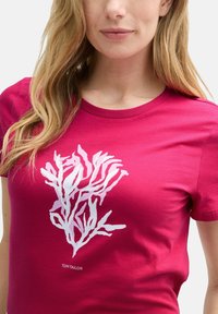 T-shirt en coton fuchsia avec un graphisme inspiré des coraux blanc sur le devant, possède un col rond classique et des manches courtes.