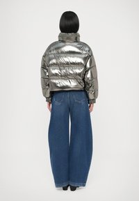 Metālisks pūkains džaka ar augstu apkakli un diagonālu šūto rakstu, kombinēts ar plašām zilām džinsu biksēm, skatīts no aizmugures.