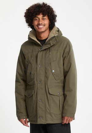 Homme aux cheveux bouclés souriant, portant une veste d'hiver à capuche vert olive avec plusieurs poches avant et des boutons-pression.