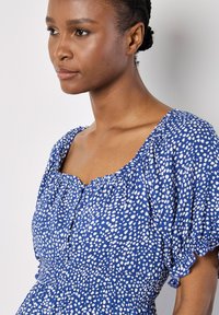 Vestido azul con mangas cortas abullonadas, con cintura fruncida y patrón de lunares blancos. Escote cuadrado con detalles de botones.
