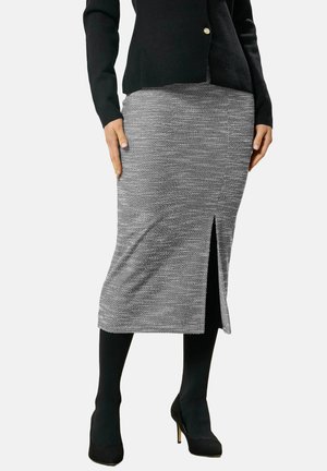 Kvinde iført en grå strikket midi-nederdel med sidelomme, sorte strømpebukser, sorte højhælede sko og en sort cardigan med knapper i guld.