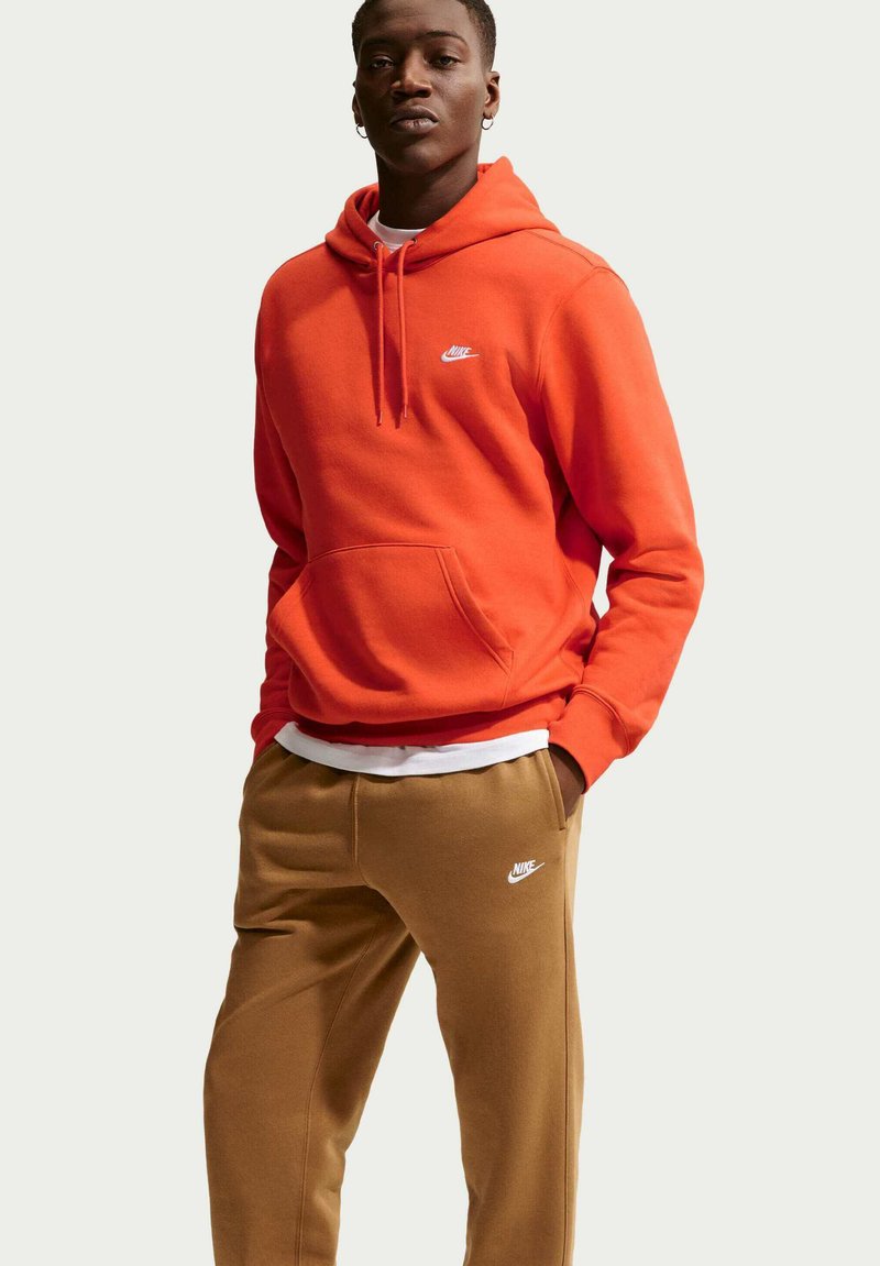 Sudadera con capucha Nike en naranja brillante con bolsillo frontal, capucha con cordón y logo blanco; combinada con pantalones deportivos marrones con logo blanco.