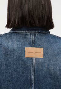 Personne aux cheveux noirs et raides vue de dos, portant une veste en jean bleue avec une pièce en cuir marron cousue sur le col arrière.