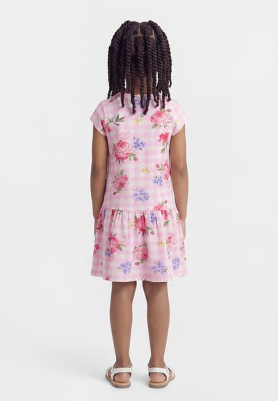 Παιδί με πλεκτά μαλλιά που φοράει ροζ καρό φόρεμα με λουλούδια και λευκά σανδάλια, στέκεται με την πλάτη προς τα εμπρός σε απλό φόντο.