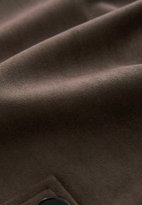 Ruda medžiaga su minkšta, dirbtine zomšos tekstūra. Medžiaga turi lygią paviršių ir subtilų blizgesį. Matomas juodas mygtukas.