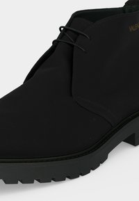 Bottine noire en cuir à lacets avec semelle épaisse crantée et détails de couture minimalistes, affichant partiellement le nom de la marque en lettres dorées.