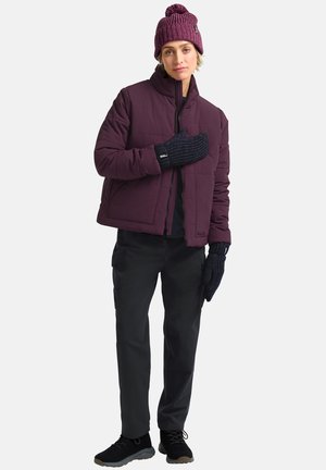 Lila gesteppte Jacke, schwarzes Oberteil, dunkle Hose, strukturierte marineblaue Handschuhe und eine gestrickte lila Mütze mit Pompon. Die Schuhe sind schwarz und haben eine strukturierte Sohle.