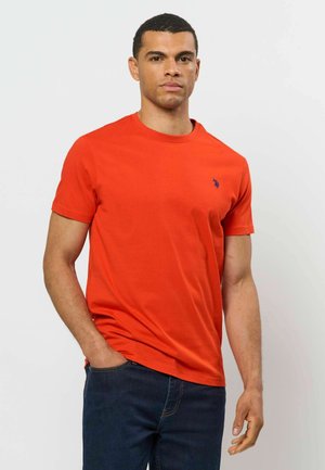 Mand stående med hånd i lommen iført en ensfarvet rød kortærmet t-shirt og mørkeblå jeans mod en lys baggrund.