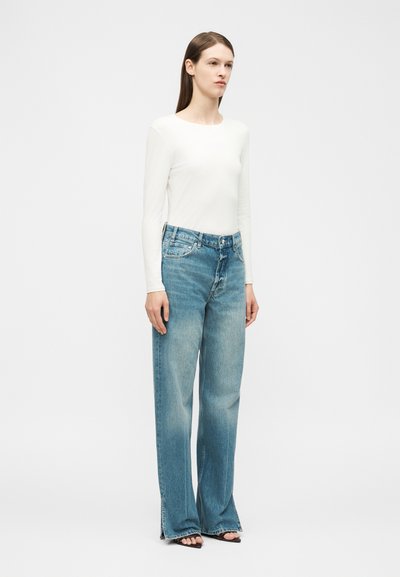 Woman standing, wearing a fitted white long-sleeve top, high-waisted wide-leg blue jeans, and black open-toe heels, plain background.