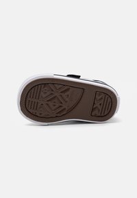 Zapatilla negra con una suela de goma texturizada marrón, entresuela blanca y una correa de velcro negra en contraste. Presenta un diseño de taco geométrico.