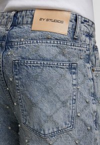 Jasnoniebieskie dżinsy z diamentowym przeszyciem i małymi ozdobami z kryształków, z brązową skórzaną naszywką z napisem "2Y STUDIOS".