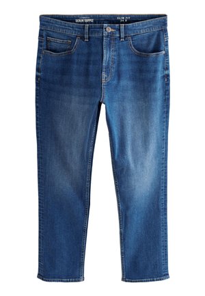 Mann trägt beige T-Shirt, beige-schwarzes kariertes Hemd, blaue Jeans und beige Sneakers, steht vor einem einfarbig hellen Hintergrund.