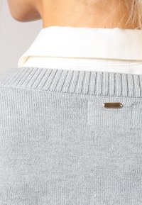 Close-up van de achterkant van een lichtgrijze gebreide trui met een klein rechthoekig metalen logo plaatje nabij de kraag, gedragen over een wit overhemd met kraag.