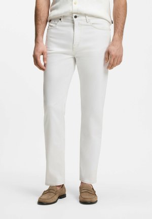 Man draagt slanke witte spijkerbroek, beige loafers en een gedeeltelijk zichtbaar wit overhemd tegen een effen achtergrond.