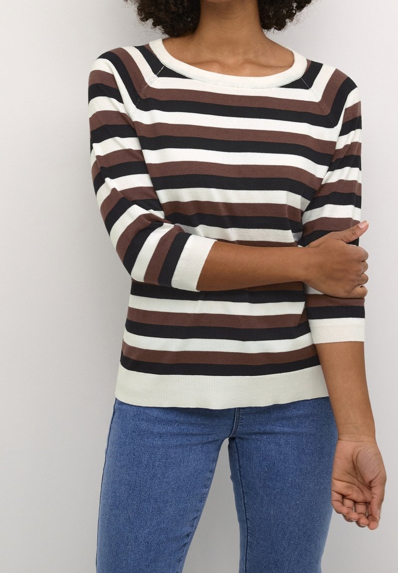 Personne portant une chemise à manches longues rayée marron, noire et blanche avec un jean bleu, tenant le bras gauche avec la main droite sur un fond clair.